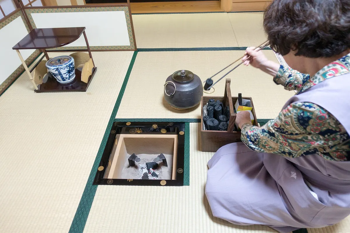 A woman in a lilac kimono kneels on a tatami floor beside a sunken hearth, lifting charcoal with long chopsticks, with a kettle and a lacquer tray of charcoal beside her