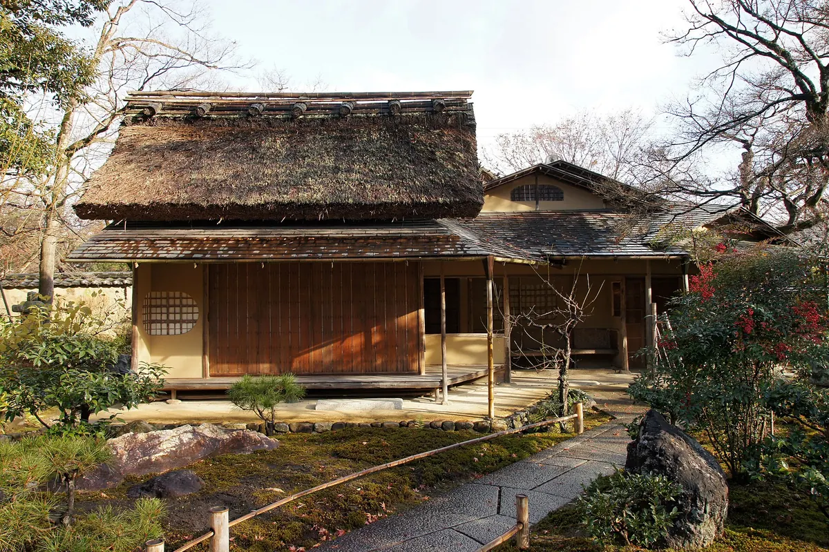 A thatched teahouse with a wooden veranda and shuttered doors in a garden with maples, a stone path and rocks, at Hogon-in temple in Kyoto