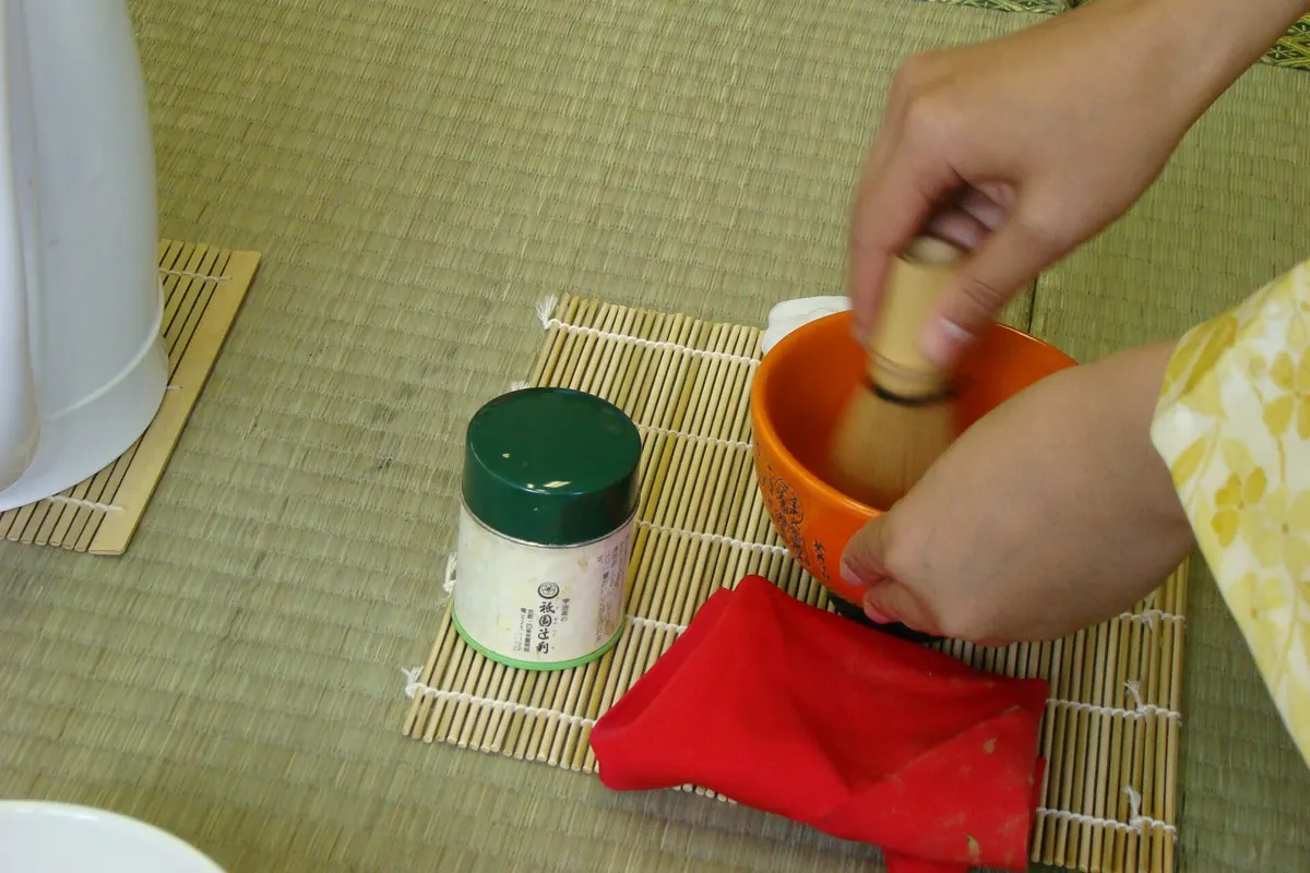 A hand whisking green tea in an orange bowl on a woven mat, with a green tea caddy and a red cloth beside it