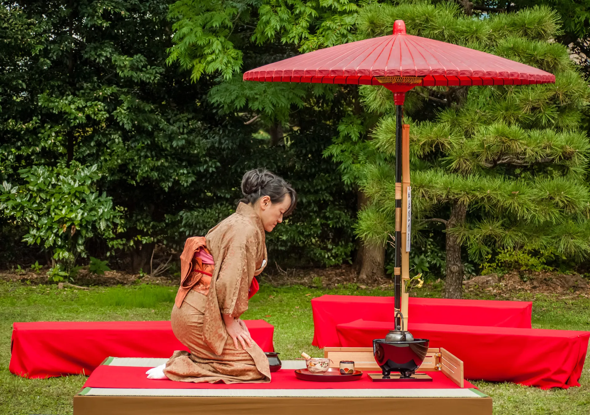 A woman in a pale brown kimono kneels at a low table set with tea utensils under a red parasol, with red benches on either side, in an outdoor tea setting in a Tokyo garden