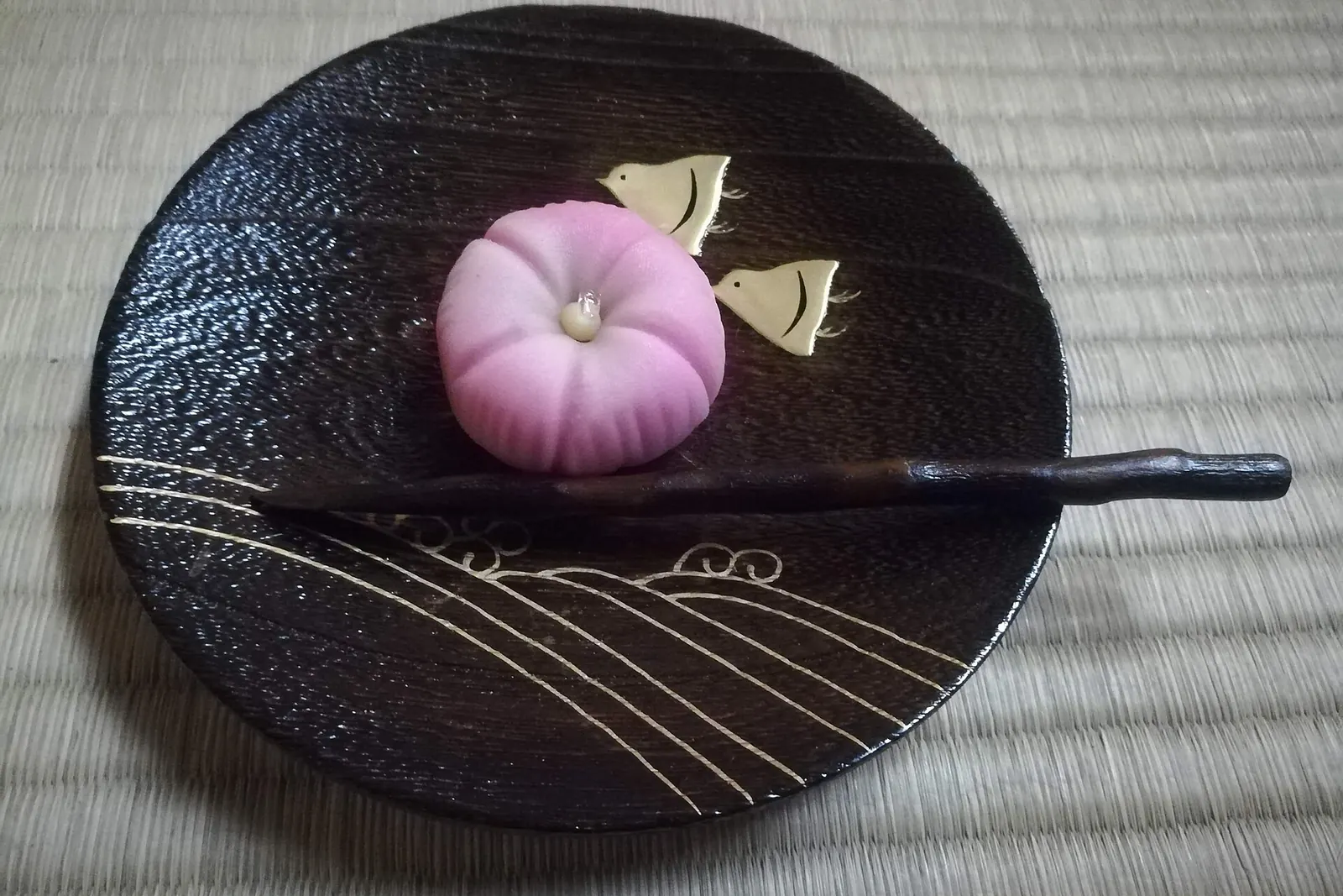 A traditional sweet, a general photograph: a pink flower-shaped confection with two small metal birds on a black lacquer plate on a tatami mat